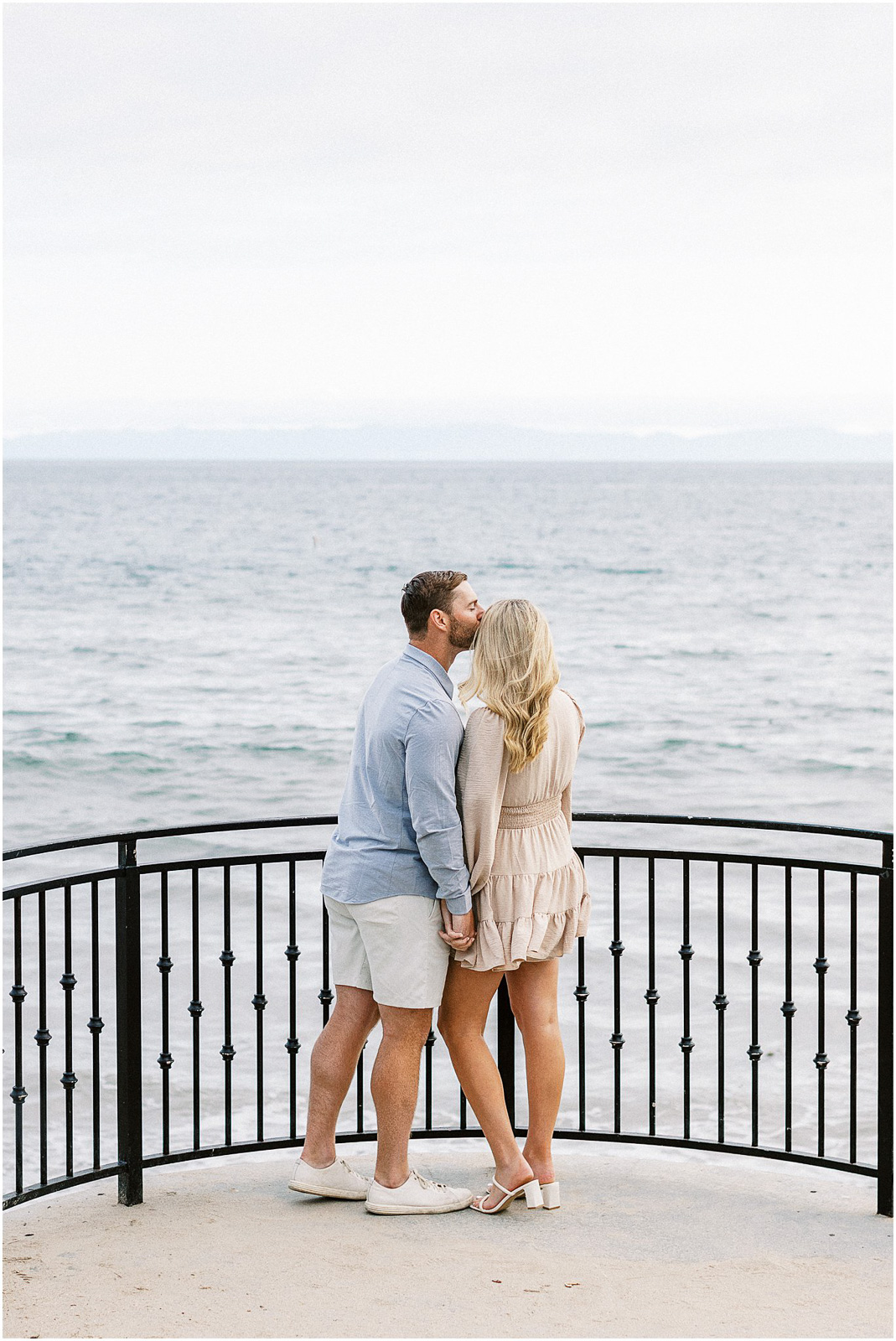 Husband kissing wife's temple in front of the pacific ocean.  