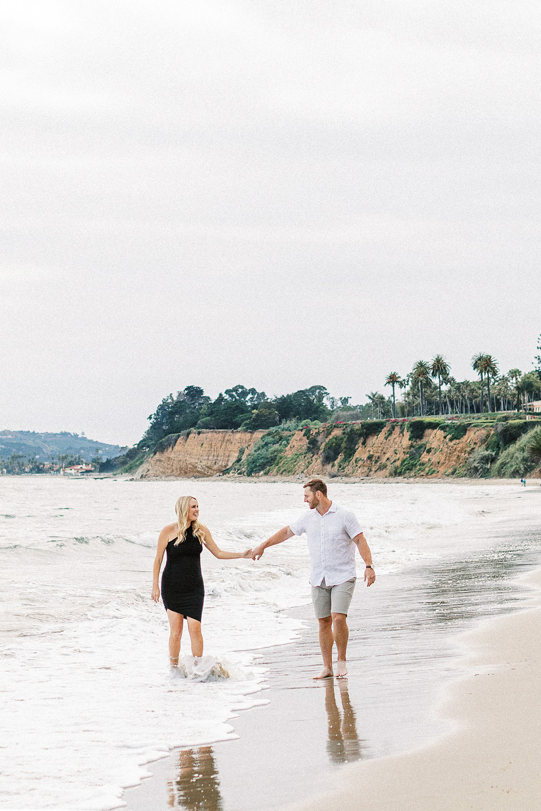 A man and woman holding hands walking in water along the beach in Montecito, Ca.