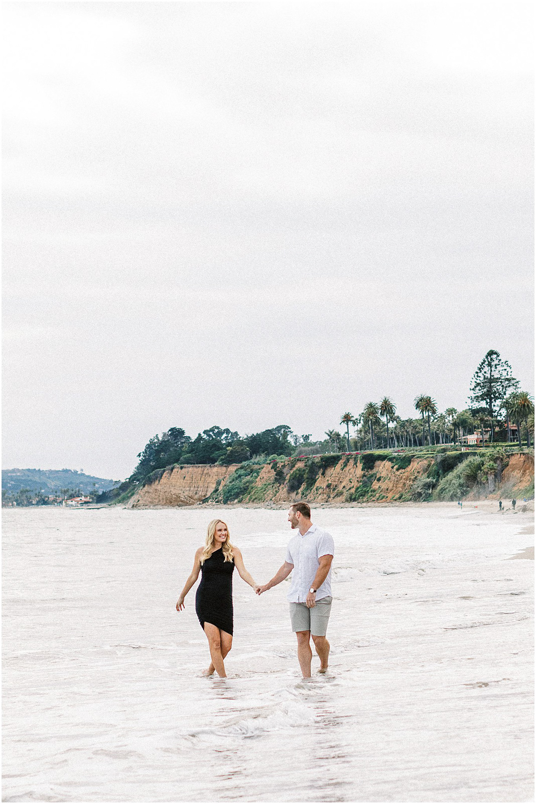 A man and woman holding hands being playful in water along the beach in Montecito, Ca.