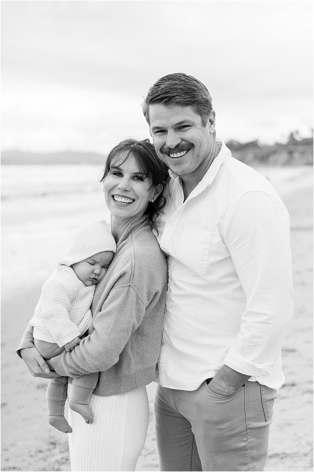 New parents smiling while holding their baby at a foggy SoCal beach