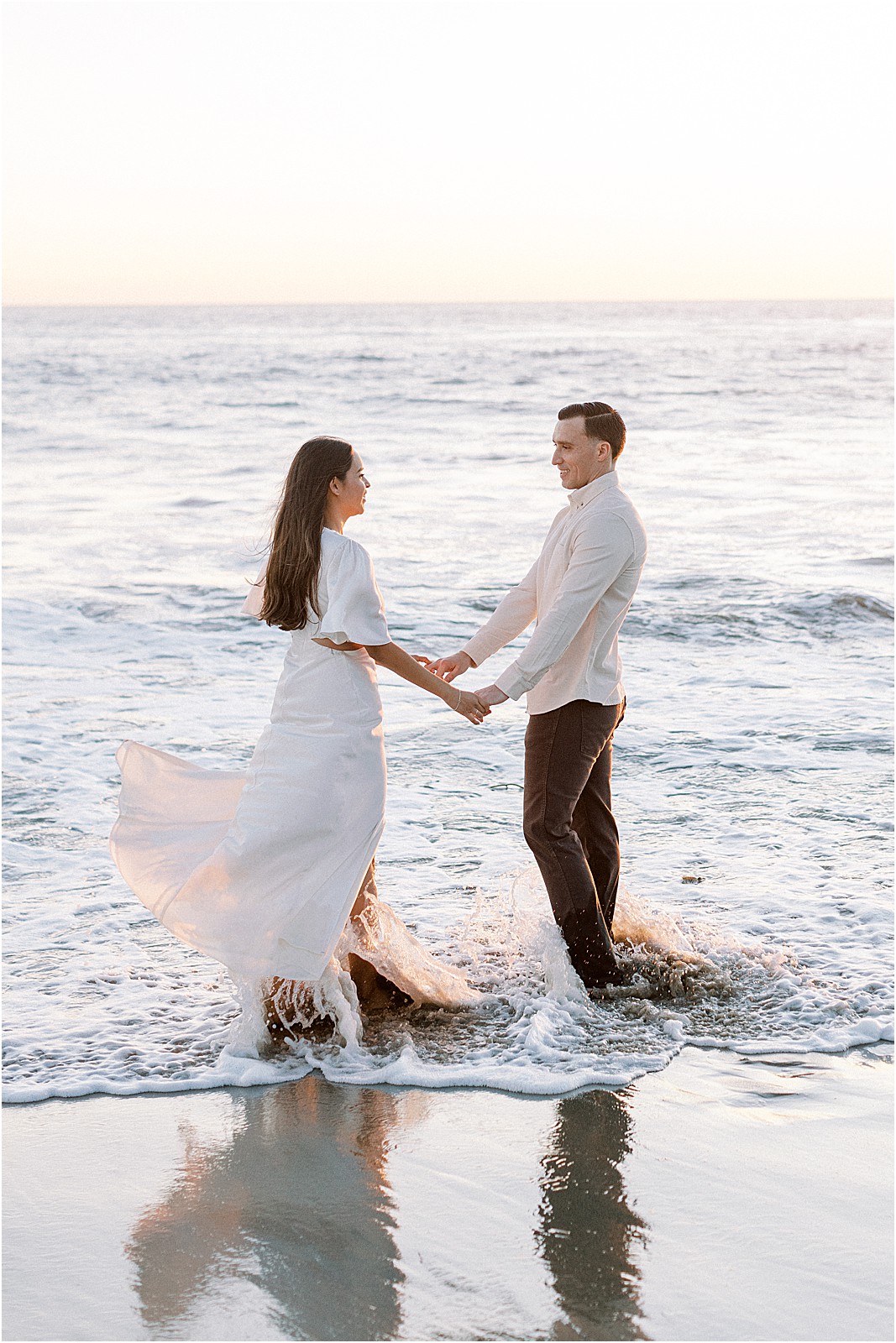 Couple holding hands while standing in the ocean at their engagement session.