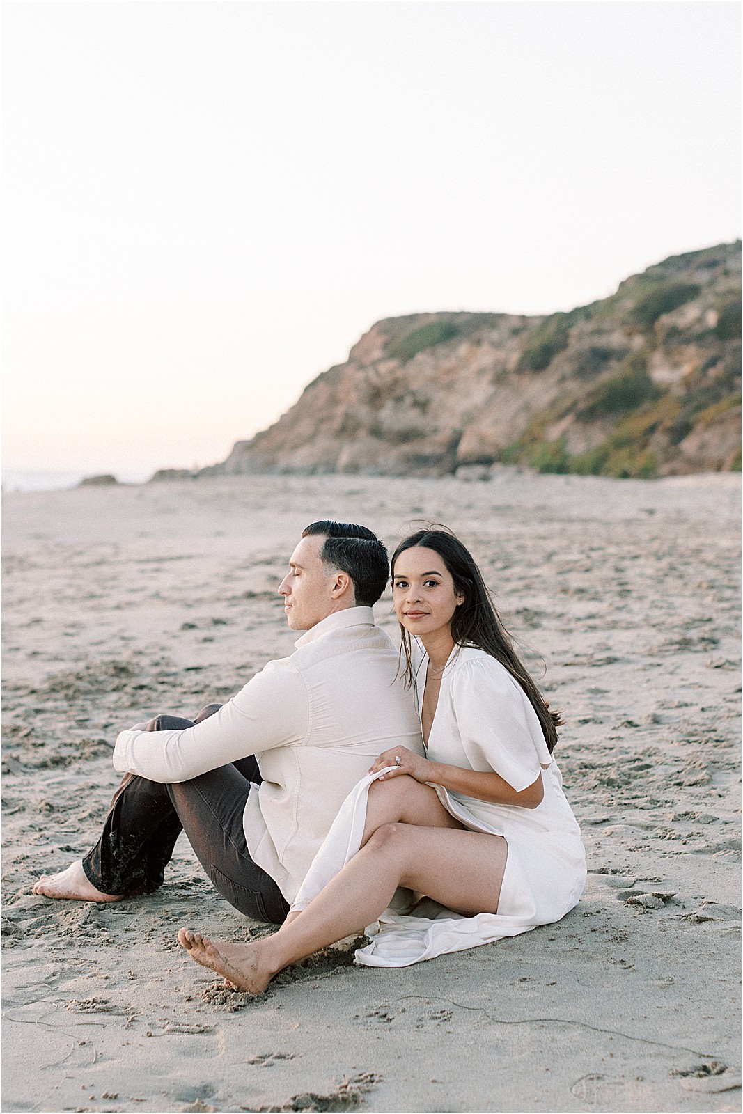 Bride smiling at the camera while sitting with groom on the sand during engagement photos in Malibu