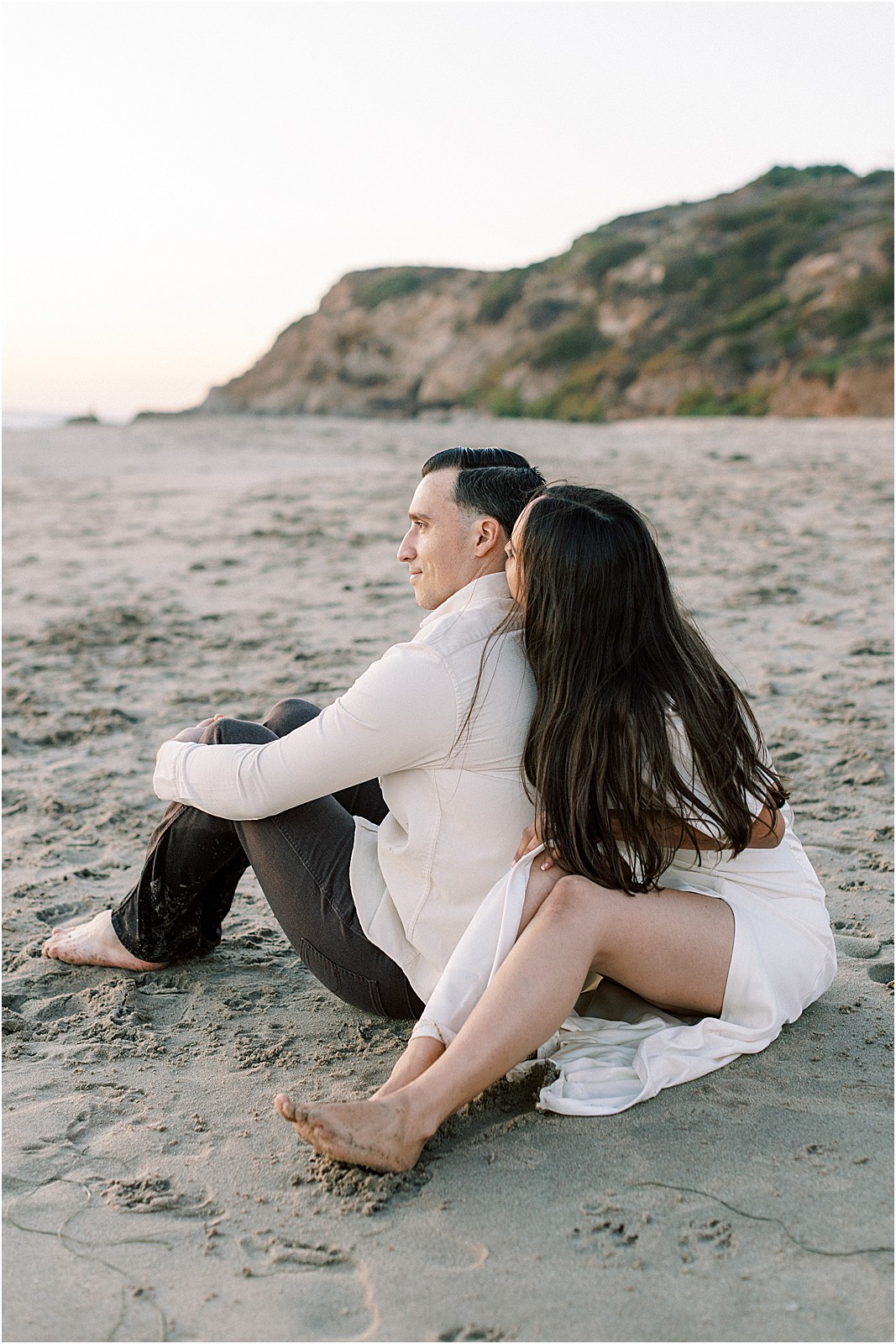 Bride leaning on groom while sitting on the beach.
