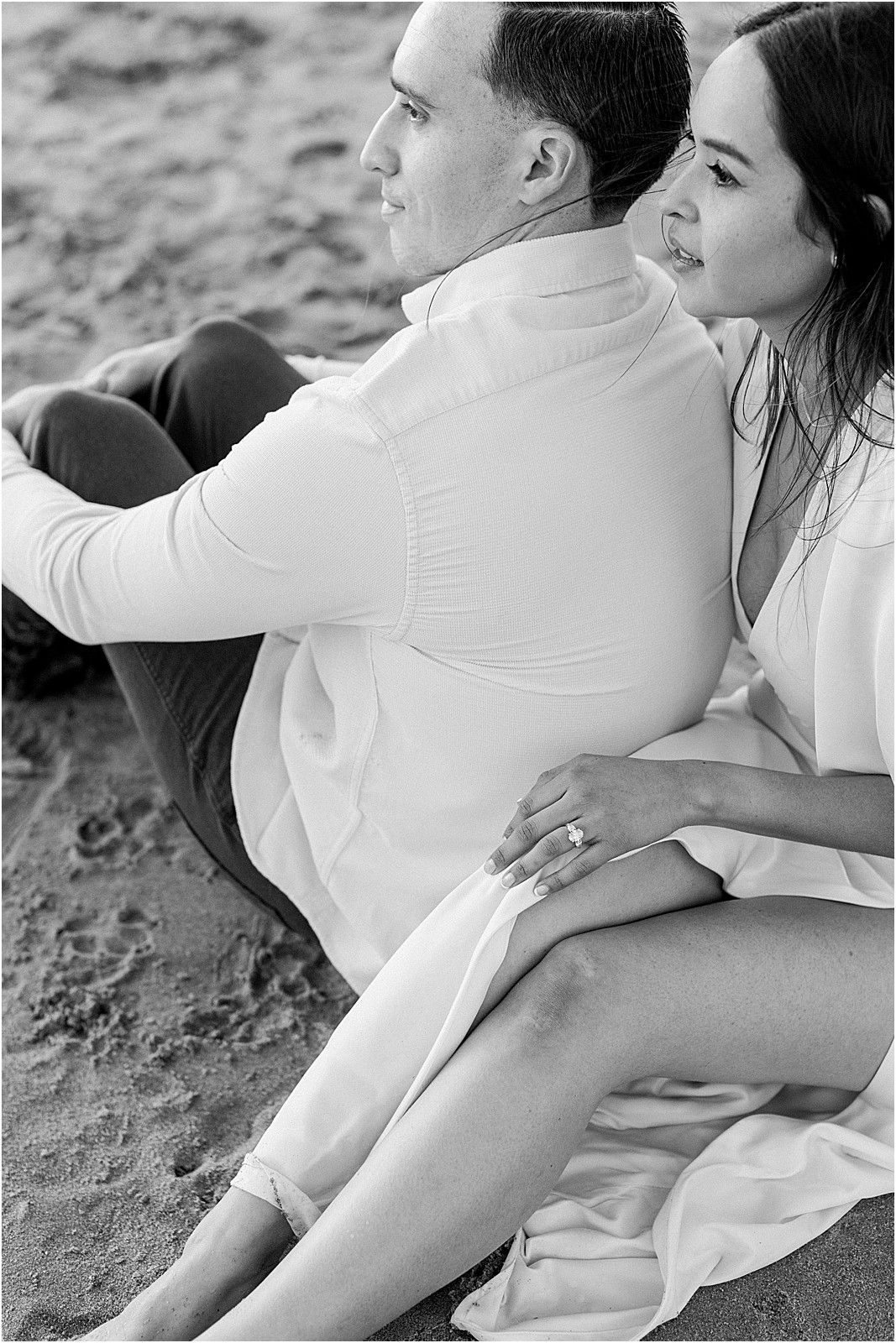 Couple sitting together on the sand showcasing her engagement ring.